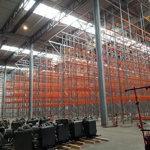 Lagerhalle mit hohen Regalen, orangefarbenen Stützen und Maschinen am Boden.