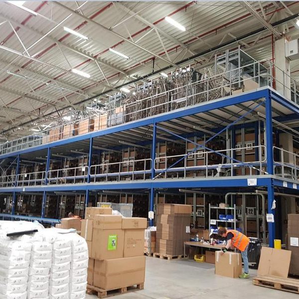 Blick auf ein Lager mit Regalen, Paletten und einem Mitarbeiter am Arbeitsplatz.