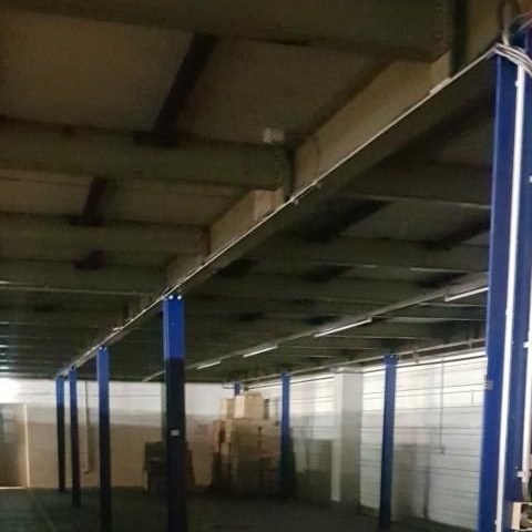 Leeres Lager mit hohen Decken und blauen Stützpfeilern. Kisten im Hintergrund.