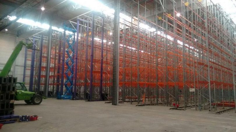 Leere Lagerhalle mit hohen Regalen in orange und blau, darunter ein Gabelstapler.