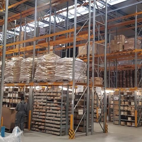 Lagerhalle mit hohen Regalen, gefüllt mit Kisten und Säcken, und einem Arbeiter im Vordergrund.