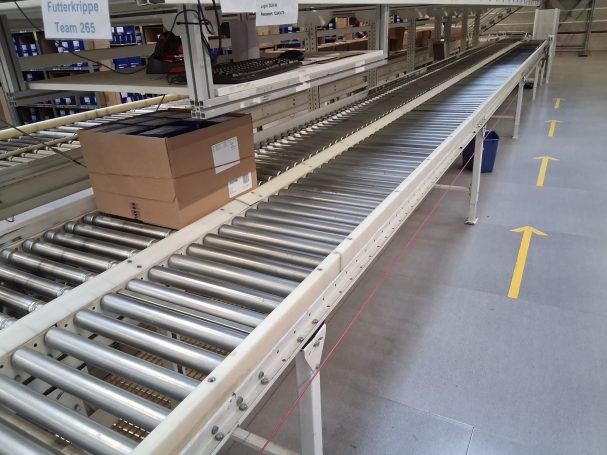 Lange Förderband mit einer Kiste darauf, in einer Lagerumgebung.