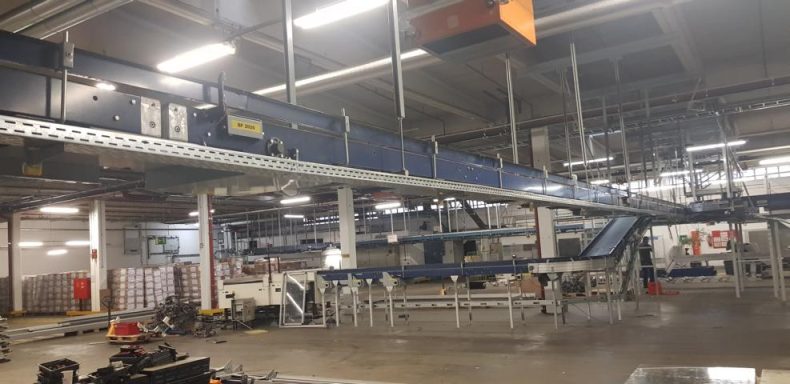 Lange Förderbandanlage in einer großen Lagerhalle mit vielen Regalen.