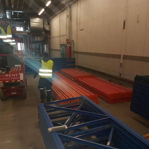 Ein Lagerraum mit orangefarbenen und blauen Regalsystemen, einer Person mit Warnweste.