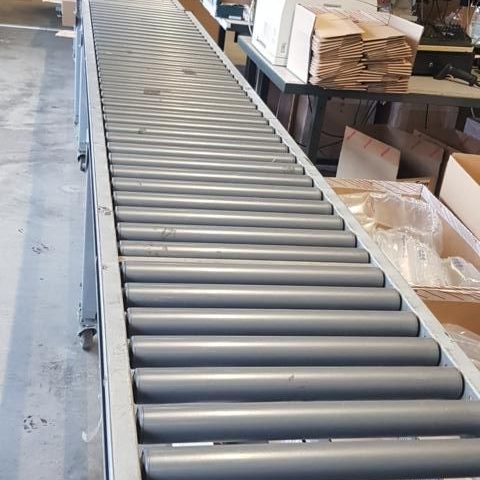 Lange Förderband mit grauen Rollen in einem Lagerraum.