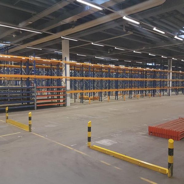 Leerer Lagerraum mit hohen Regalen in orange und blau sowie gelben Abgrenzungen.