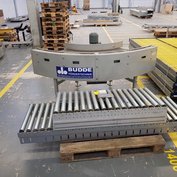 Transportband mit Rollen im Lager, umgeben von Paletten und weiteren Geräten.