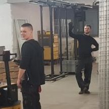 Zwei Männern in Arbeitskleidung in einem Lager mit Regalen und einem Gabelstapler.