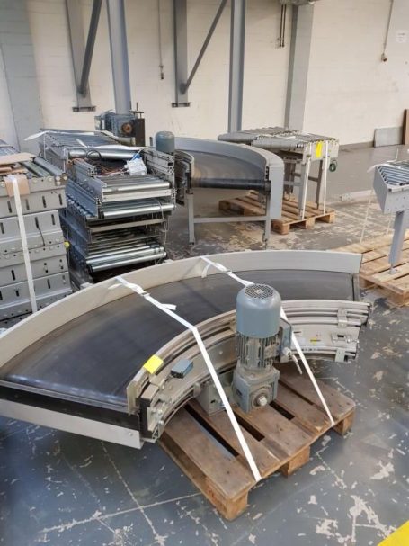 Krummförmige Förderbänder und Maschinen auf Holzpaletten in einer Lagerumgebung.