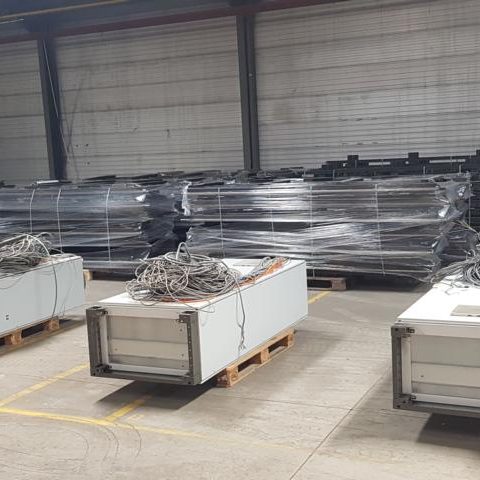 Lagerraum mit gestapelten Containern und Metallteilen auf Paletten.