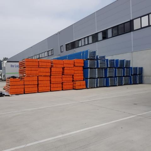 Stapelhaufen aus orangefarbenen und blauen Containern vor einer Lagerhalle.