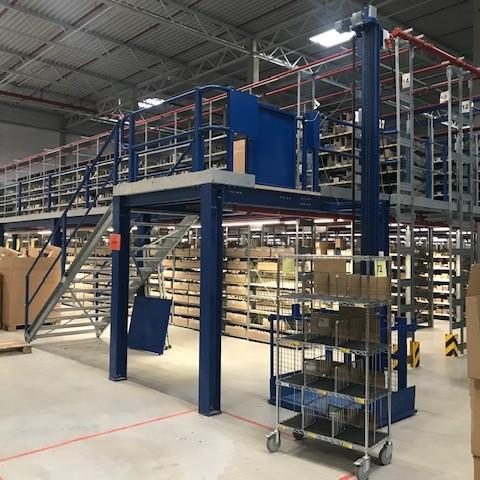 Regalstruktur in einem Lager mit Treppe und Transportwagen im Vordergrund.