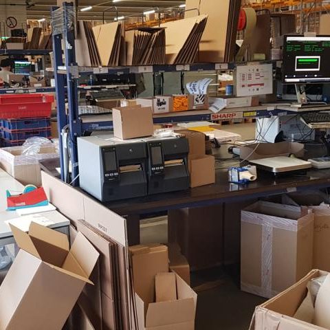 Lagerraum mit Kartons, Verpackungsmaschinen und Arbeitstisch.