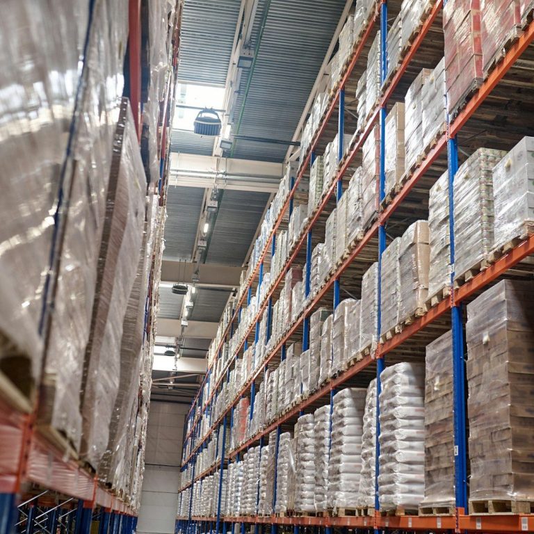 Lagerhalle mit hohen Regalen, voll mit Paletten und Verpackungen.