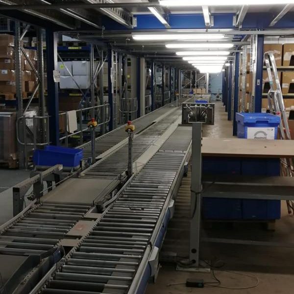 Lange Förderbandanlage in einem Lager, mit Regalen und Arbeitsplätzen im Hintergrund.