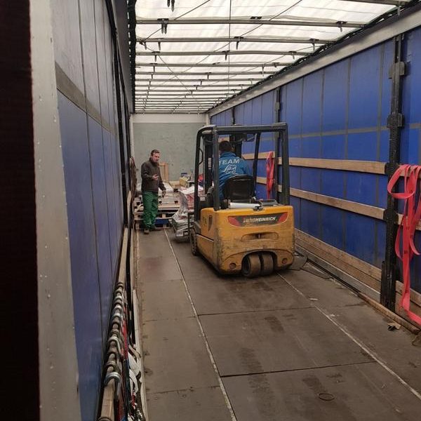 Gabelstapler transportiert Waren in einem Lkw, zwei Personen arbeiten im Hintergrund.
