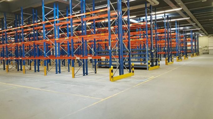 Lagerhalle mit blauen Regalen und orangefarbenen Trägersystemen, leere Flächen.
