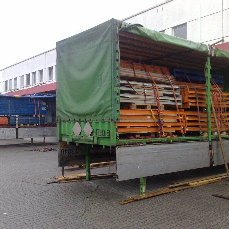 Lkw mit gestapelten Holzpaletten in orange und blau, vor einem Gebäude.