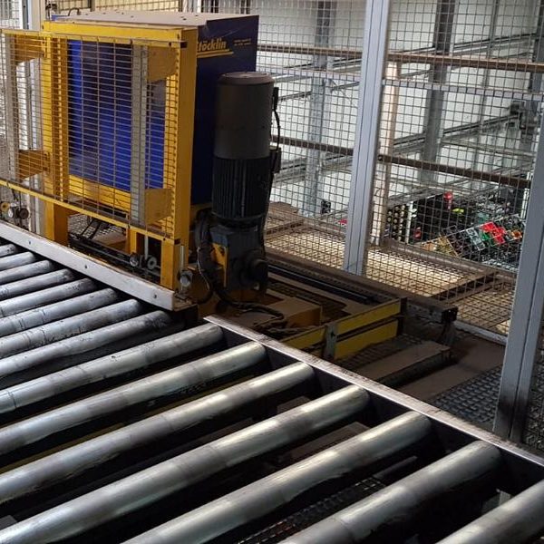 Förderband mit gelber Maschinenstation im Hintergrund, umgeben von Metallgittern.