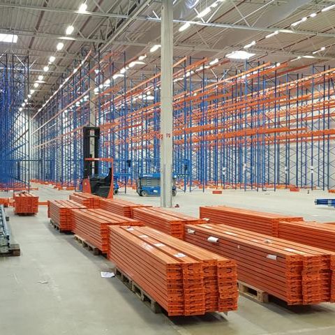 Lagerhalle mit blauen und orangen Regalen sowie gestapelten Holzlatten.