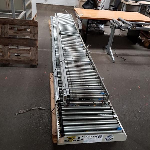 Lange Förderbandanlage aus Metall mit mehreren Rollen auf Holzunterlage.