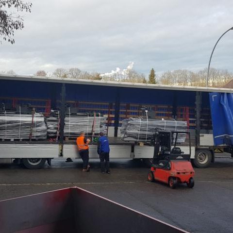 Lkw mit mehreren Paletten, zwei Personen stehen daneben, Gabelstapler in der Nähe.