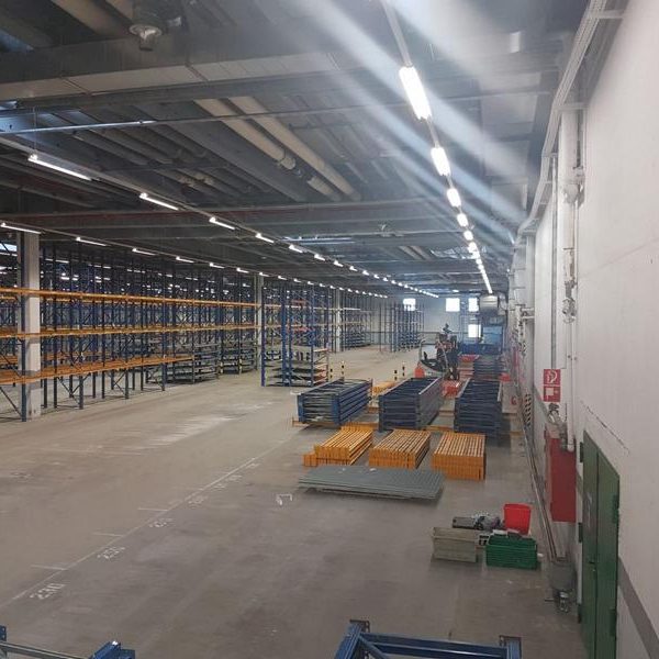 Lagerhalle mit Regalen, leeren Bereichen und bunten Paletten auf dem Boden.