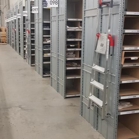 Regale mit leeren Fächern in einem Lagerraum, helle Wände, grauer Boden.