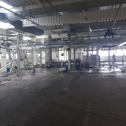 Leere Industriehalle mit Maschinen und großen Fenstern, schwach beleuchtet.