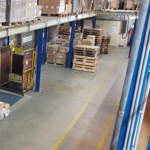 Lagerhalle mit Holzpaletten und Regalen, geräumiger Boden und gelbe Markierungen.