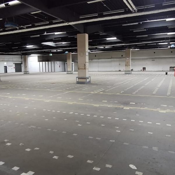 Leere Halle mit grauem Boden und sparsamer Einrichtung, Säulen in der Mitte.