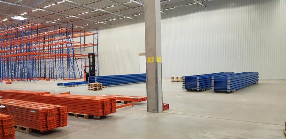 Lagerbereich mit orangefarbenen und blauen Regalen, leerem Raum und Gabelstapler.